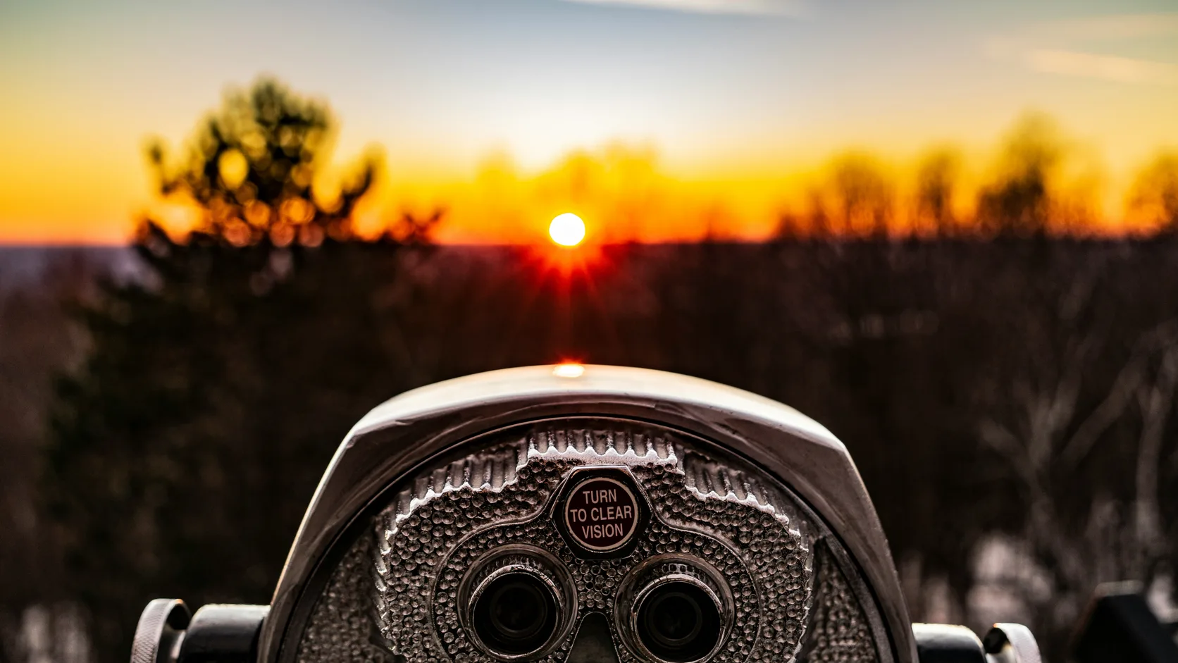 A coin-operated viewing telescope pointed at the sun setting over a tree line