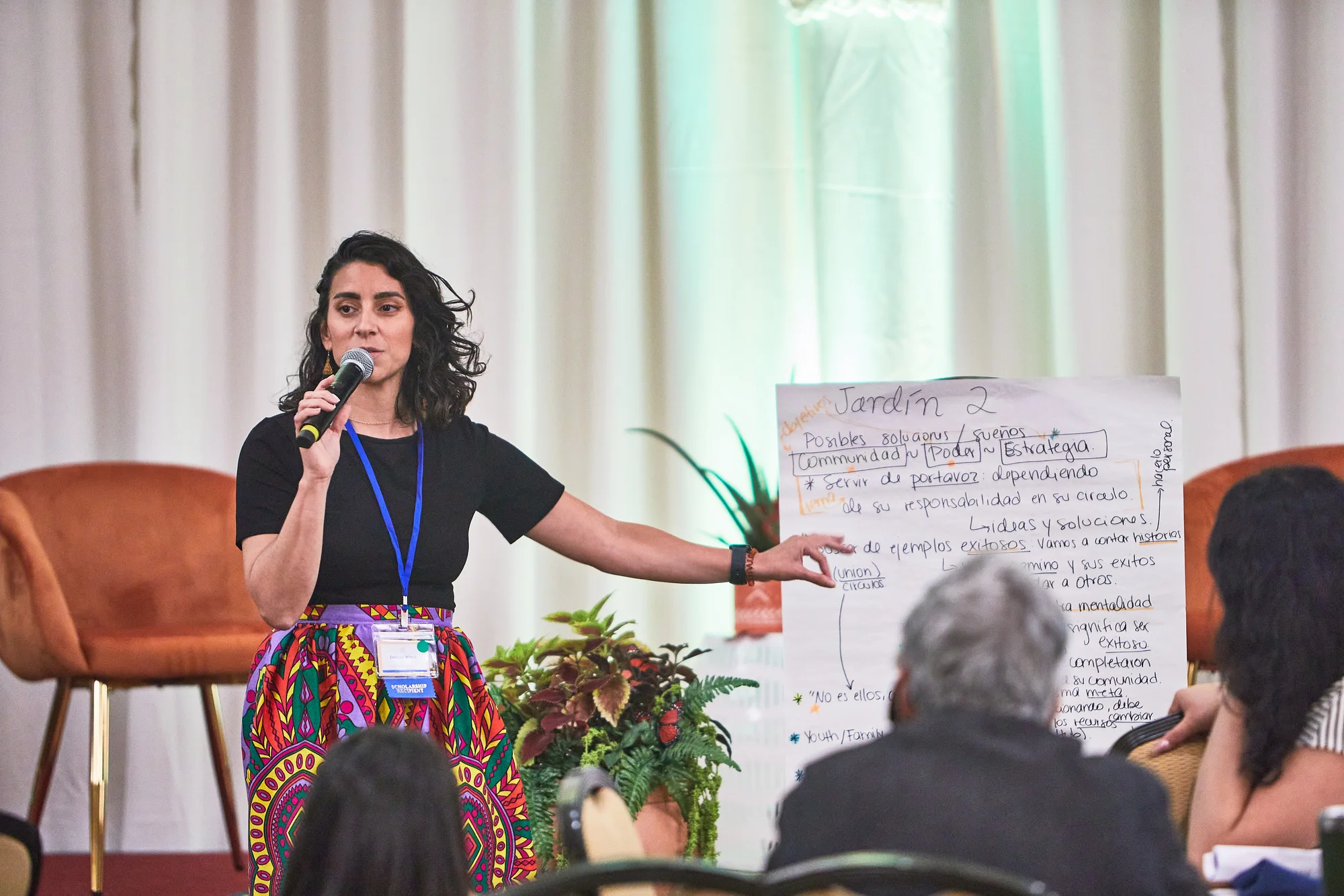 A LatinxEd leader speaks into a microphone in front of a workshop poster written in Spanish